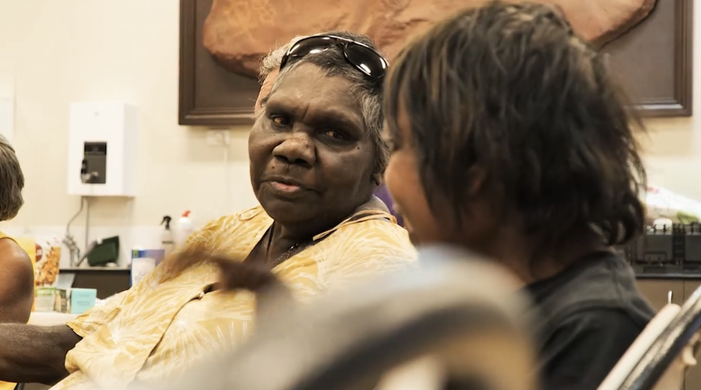 Indigenous Elder in bright yellow dress smiles at a little girl whose back is to us.